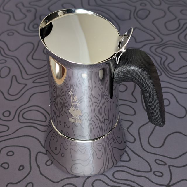 The fully assembled stainless steel moka pot standing upright on the topographic mat. Its highly polished surface distinctly reflects the wavy contour lines of the mat beneath it. It features a solid black, curved handle.