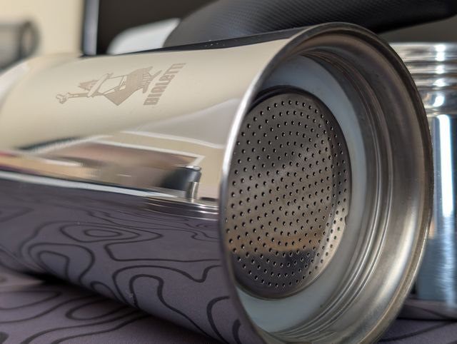 A close up of the top half of a stainless steel Bialetti Venus moka pot lying on its side on a dark grey topographic-patterned mat. The camera focuses on the metal filter plate with its grid of small holes and the surrounding rubber gasket.
